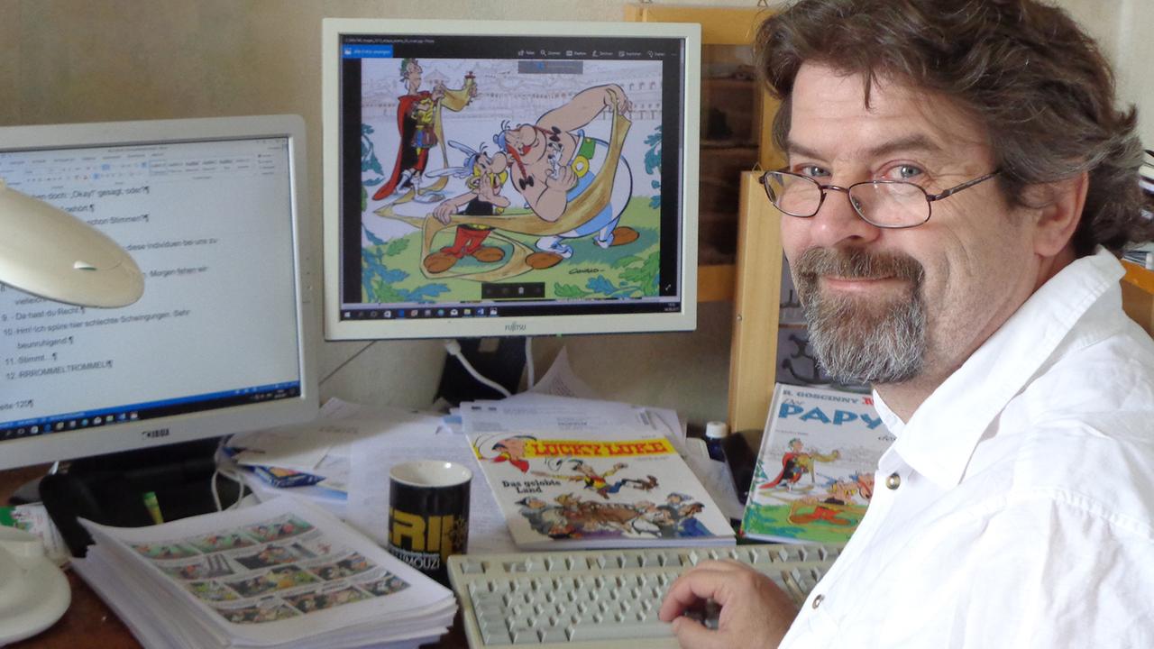 Klaus Jöken sitzt an zwei Monitoren, links ein Textdokument, rechts eine Zeichnung aus dem Comic von Asterix und Obelix.