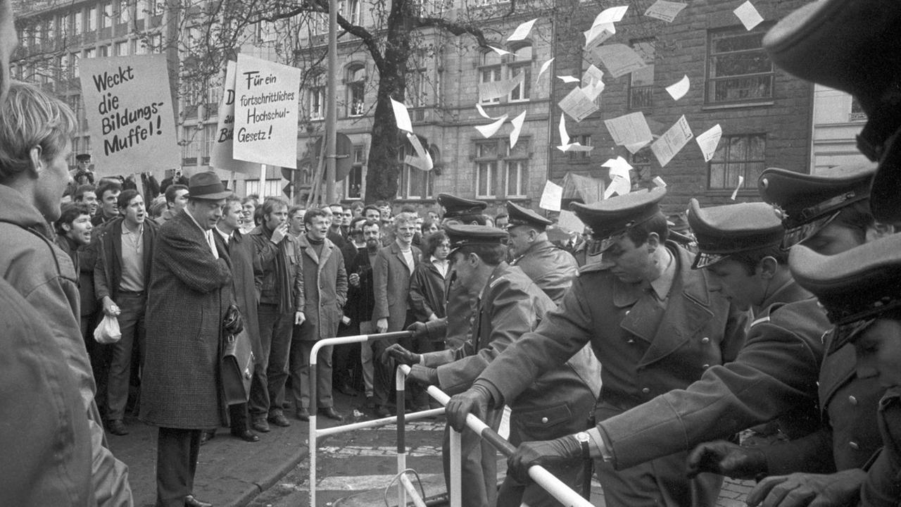 Studierende demonstrieren im April 1969 mit Plakaten und Flyern. Gegenüber stehen ihnen uniformierte Beamte hinter einem Zaun.