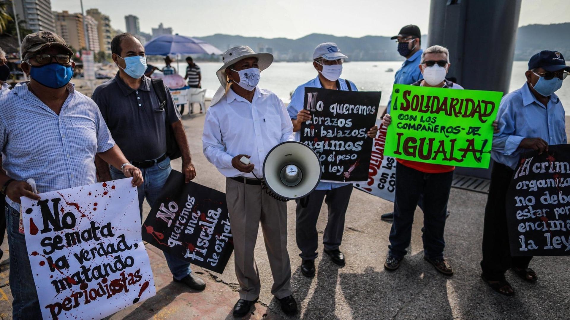Mehrere Männer mit Gesichtsmasken, einem Megafon und beschrifteten Plakaten stehen an einer Hafenmole.