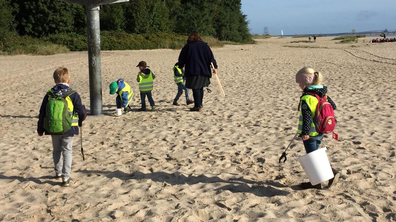 Kinder in gelben Warnwesten sammeln Müll am Strand auf. 
