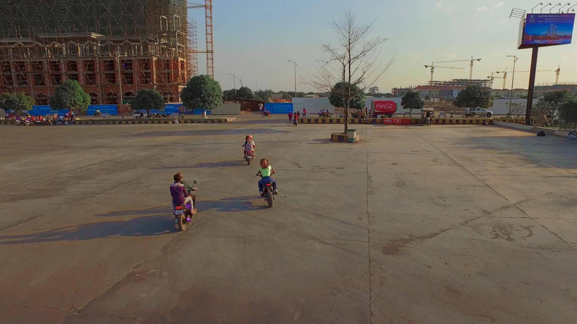In einer Szene aus Davy Chous Film "Diamond Island" ist ein großer leerer Platz zu sehen, über den drei Mofas fahren; im Hintergrund Häuserbaustellen.