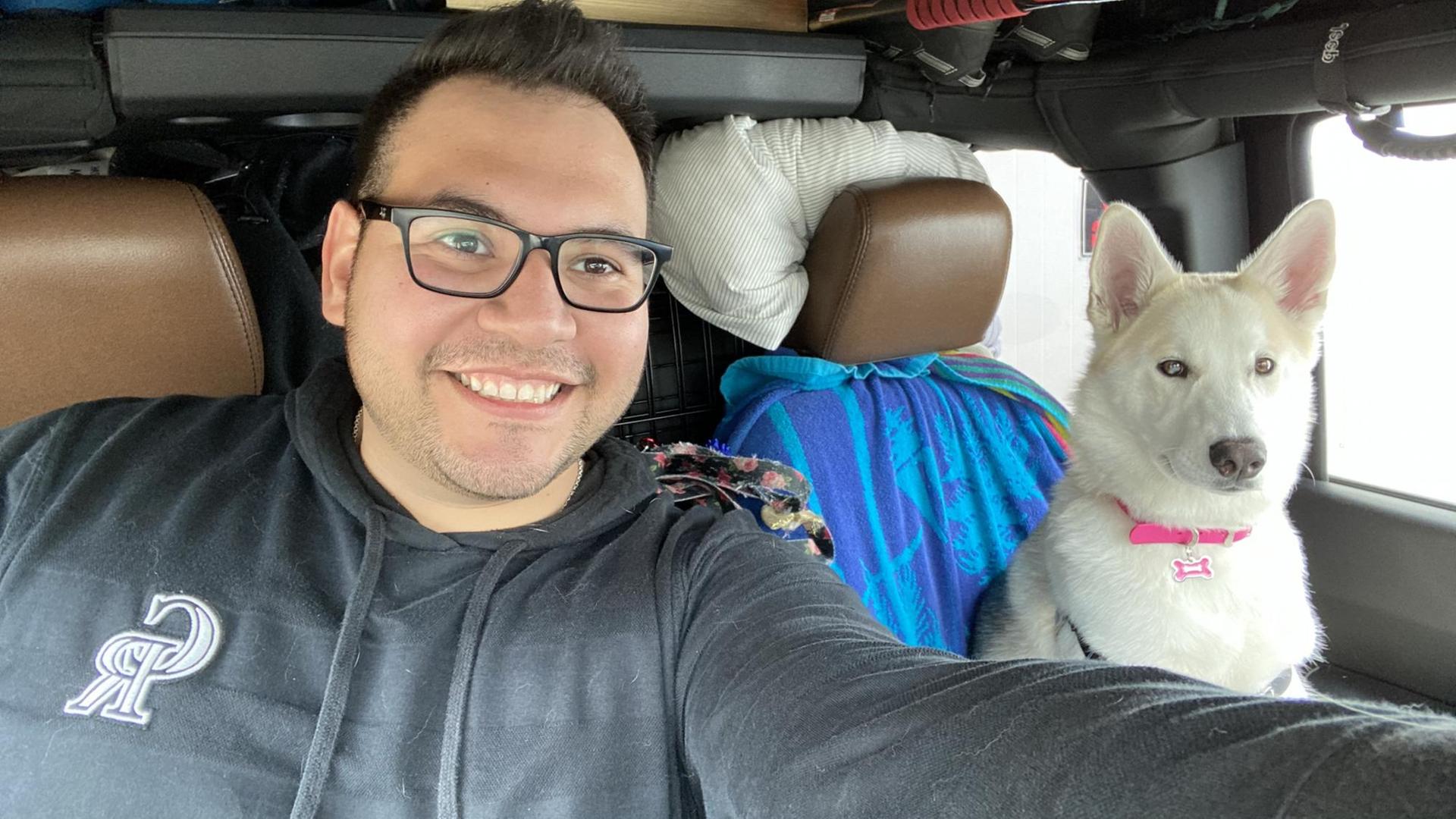 Ein lächelnder junger Mann, Oscar Garza, mit Brille sitzt im Auto. Neben ihm eine weiße Hündin auf dem Beifahrersitz.