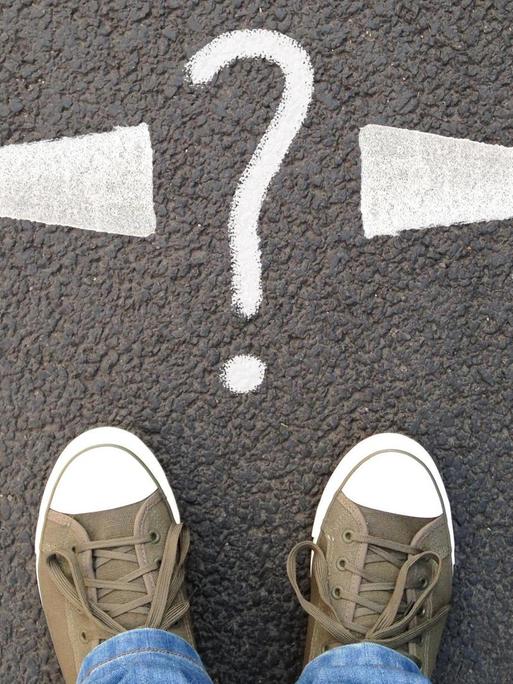 Füße in Turnschuhen stehen vor einer Markierung auf dem Asphalt: ein Fragezeichen und zwei Pfeile in entgegengesetzte Richtungen.
