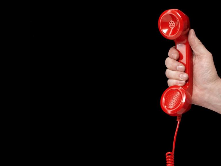 Eine Hand hält einen roten Telefonhörer vor schwarzem Hintergrund.