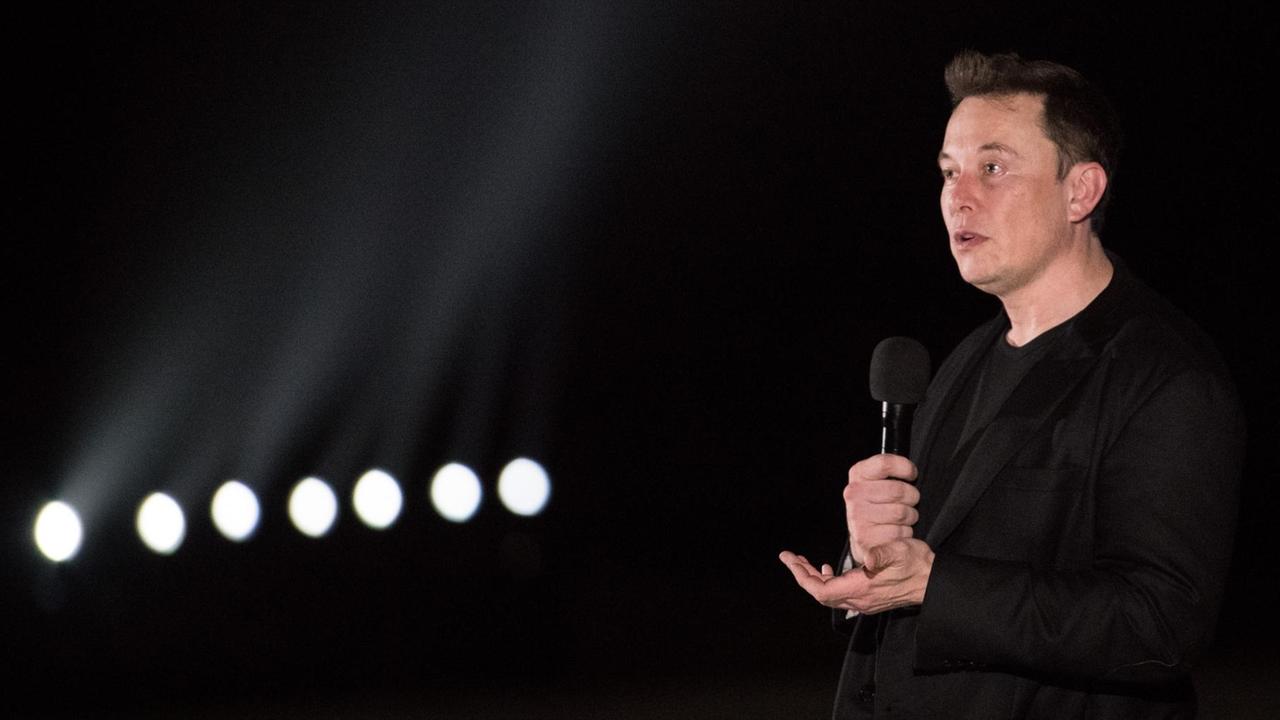 Elon Musk bei einer Rede in Texas, er steht vor dunklem Hintergrund und hält ein Mikrofon in der Hand.