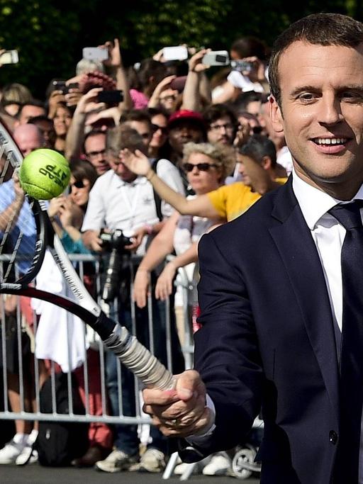 Der französische Staatspräsident Emmanuel Macron trägt Anzug und spielt Tennis.