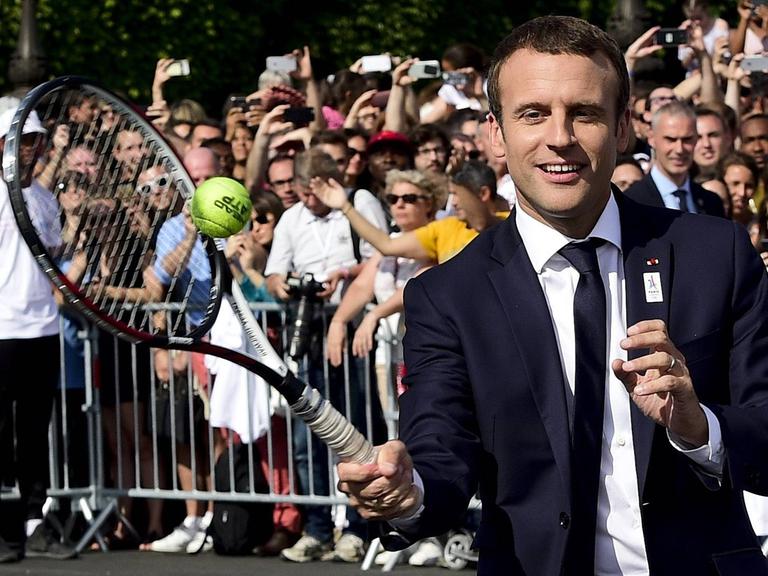 Der französische Staatspräsident Emmanuel Macron trägt Anzug und spielt Tennis.