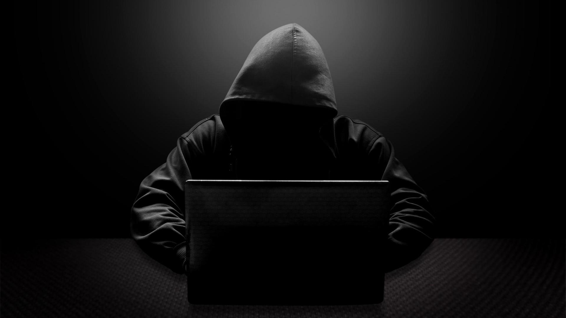 Ein Mann mit schwarzer Kapuze und verschattetem Gesicht sitzt vor einem Laptop.
