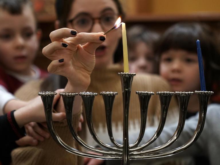 Eine Gruppe von Kindern und einer Frau zünden eine Chanukkah Kerze zum jüdischen Lichterfest am neunarmigen Leuchter an.
