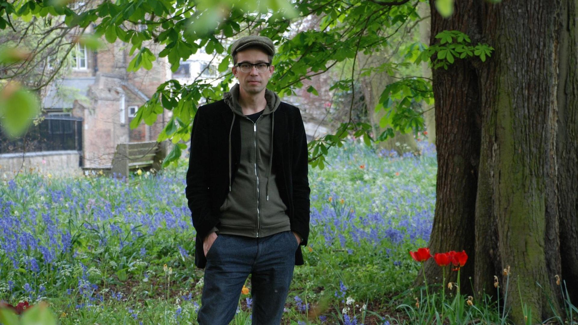 Der Historiker Sören Urbansky mit Schiebermütze und Brille steht auf einer Wiese unter einem Baum.