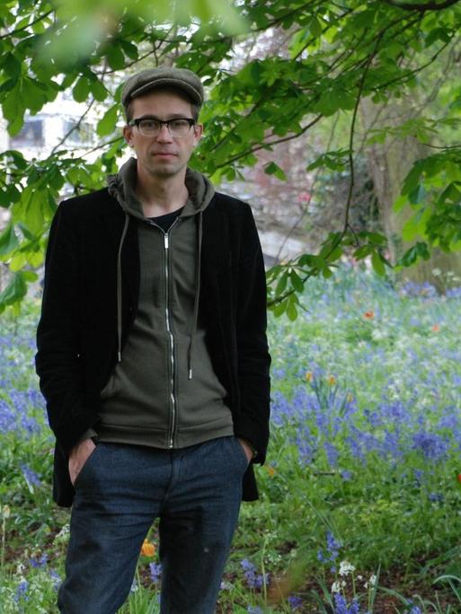 Der Historiker Sören Urbansky mit Schiebermütze und Brille steht auf einer Wiese unter einem Baum.