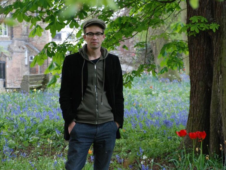 Der Historiker Sören Urbansky mit Schiebermütze und Brille steht auf einer Wiese unter einem Baum.