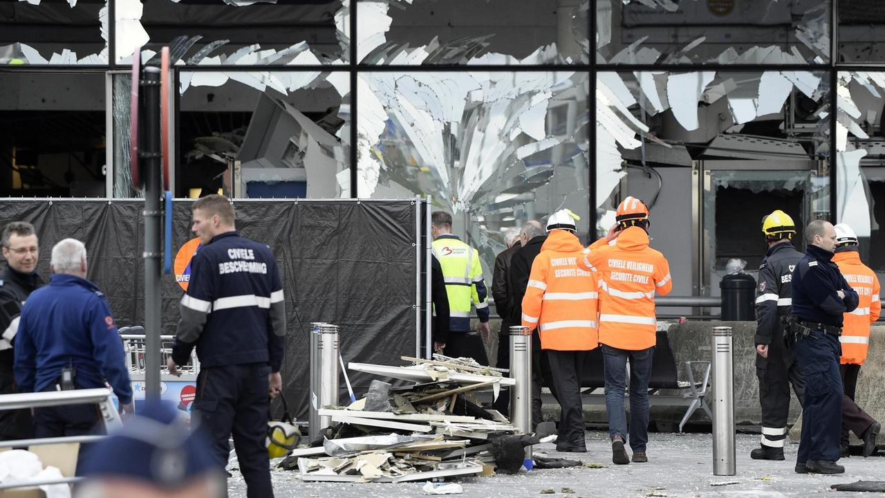 Zerborstene Scheiben am Terminal des Brüsseler Flughafens. Davor stehen Menschen mit Schutzhelmen und Warnwesten. Zerborstene Scheiben am Terminal des Brüsseler Flughafens. Davor stehen Menschen mit Schutzhelmen und Warnwesten.