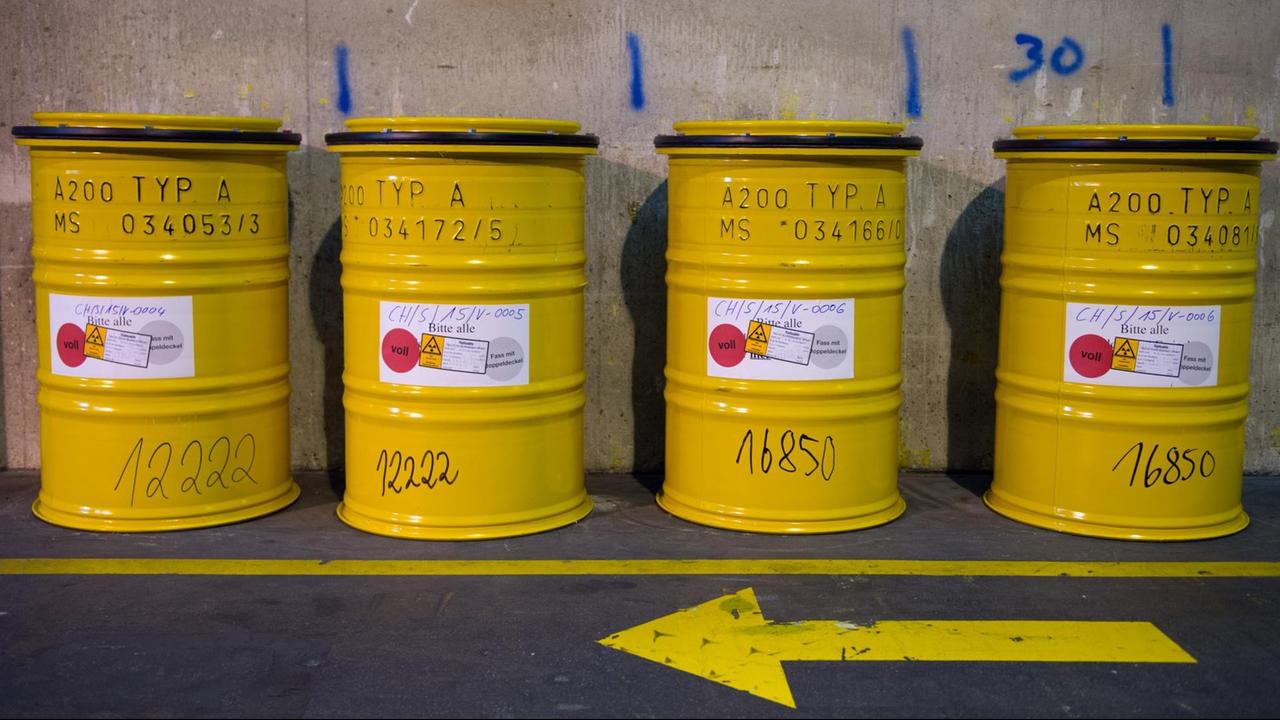 Vier gelbe Fässer mit radioaktivem Abfall stehen neben einem Weg auf dem ein gelber Richtungspfeil am Boden nach rechts zeigt.
