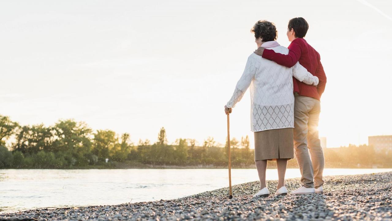 Mutter und erwachsene Tochter blicken im Abendlicht auf einen Fluss. Sie halten einander im Arm.