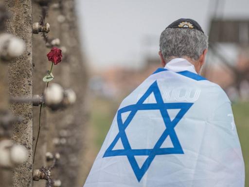 Ein in eine Israelfahne gehüllter Mann steht mit dem Rücken zur Kamera neben einer Wand im früheren Konzentrationslager Auschwitz, an der Gedenk-Devotionalien angebracht sind.