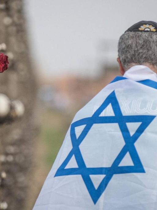 Ein in eine Israelfahne gehüllter Mann steht mit dem Rücken zur Kamera neben einer Wand im früheren Konzentrationslager Auschwitz, an der Gedenk-Devotionalien angebracht sind.