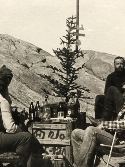 Alte Schwarzweiß-Aufnahme aus den 80er-Jahren, die zeigt, wie die Expeditionsteilnehmer in der Antarktis im Freien um eine als Tisch dienende Holzkiste herumsitzen. Darauf Flaschen und ein karger Weihnachtsbaum mit ein paar Christbaumkugeln.