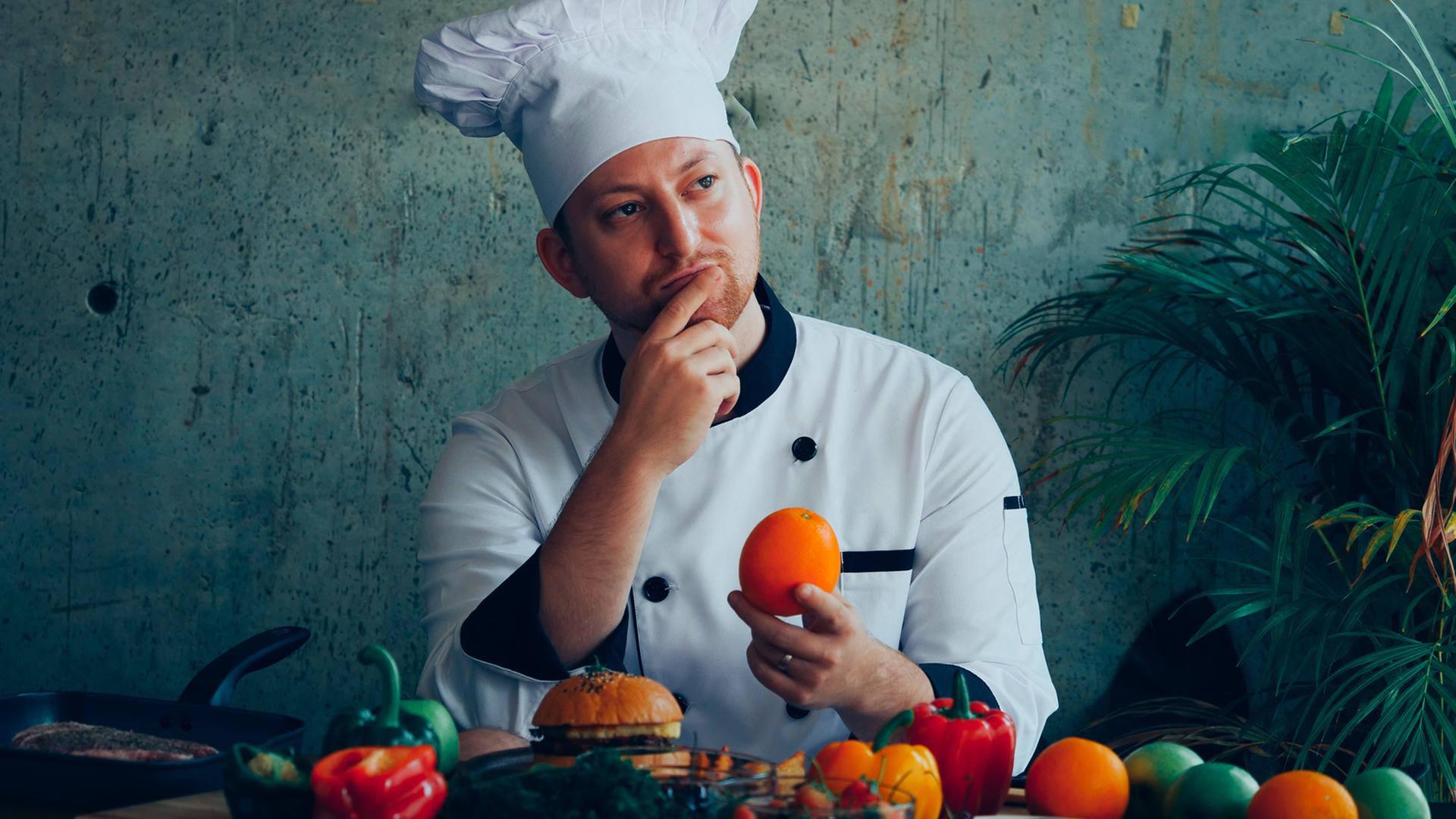 Ein junge Mann mit einer Kochmütze sitzt an einem Tisch, vor sich Obst und Gemüse. In der einen Hand hält er eine Orange, die andere Hand liegt an seinem Kinn.
