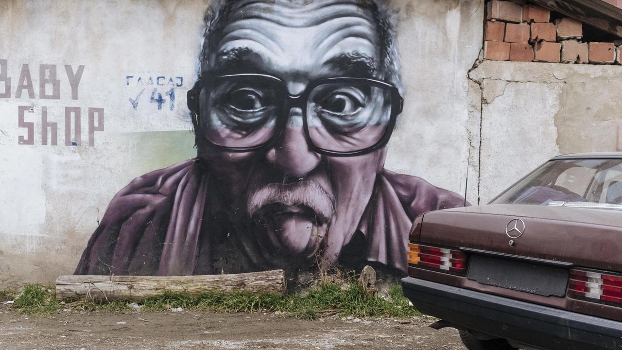 Ein Auto ohne Nummernschild steht vor einer Wand mit einem Street-Art-Porträt eines Manes, der die Zunge rausstreckt

