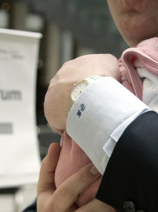 Ein Mann im Anzug trägt ein kleines Baby auf dem Arm, im Hintergrund ist ein Schild mit der Aufschrift "Tagungszentrum" zu sehen.