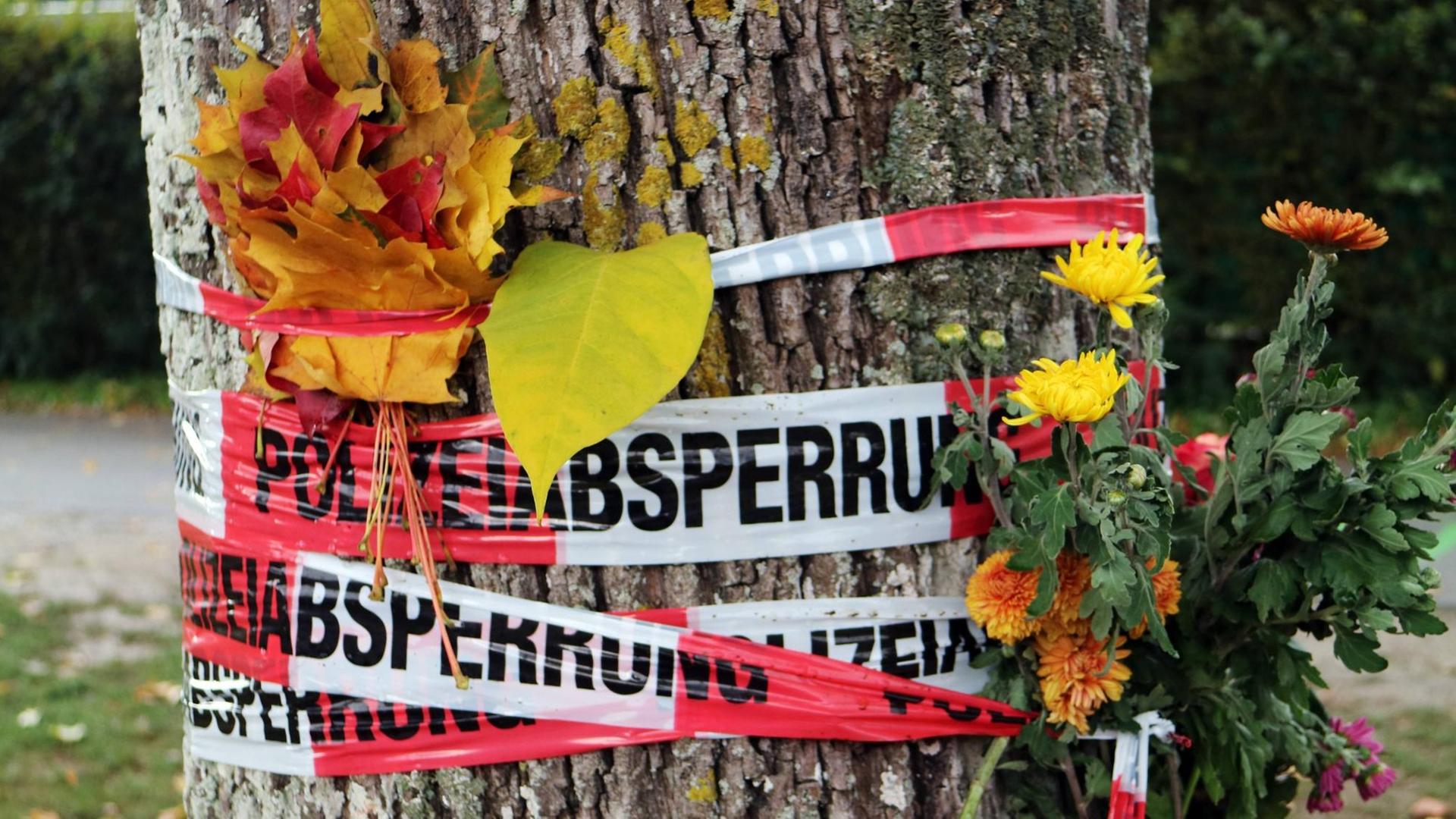Blumen und Herbstlaub stecken zwischen den Absperrbändern der Polizei an der Dreisam in Freiburg, wo eine junge Frau getötet wurde.