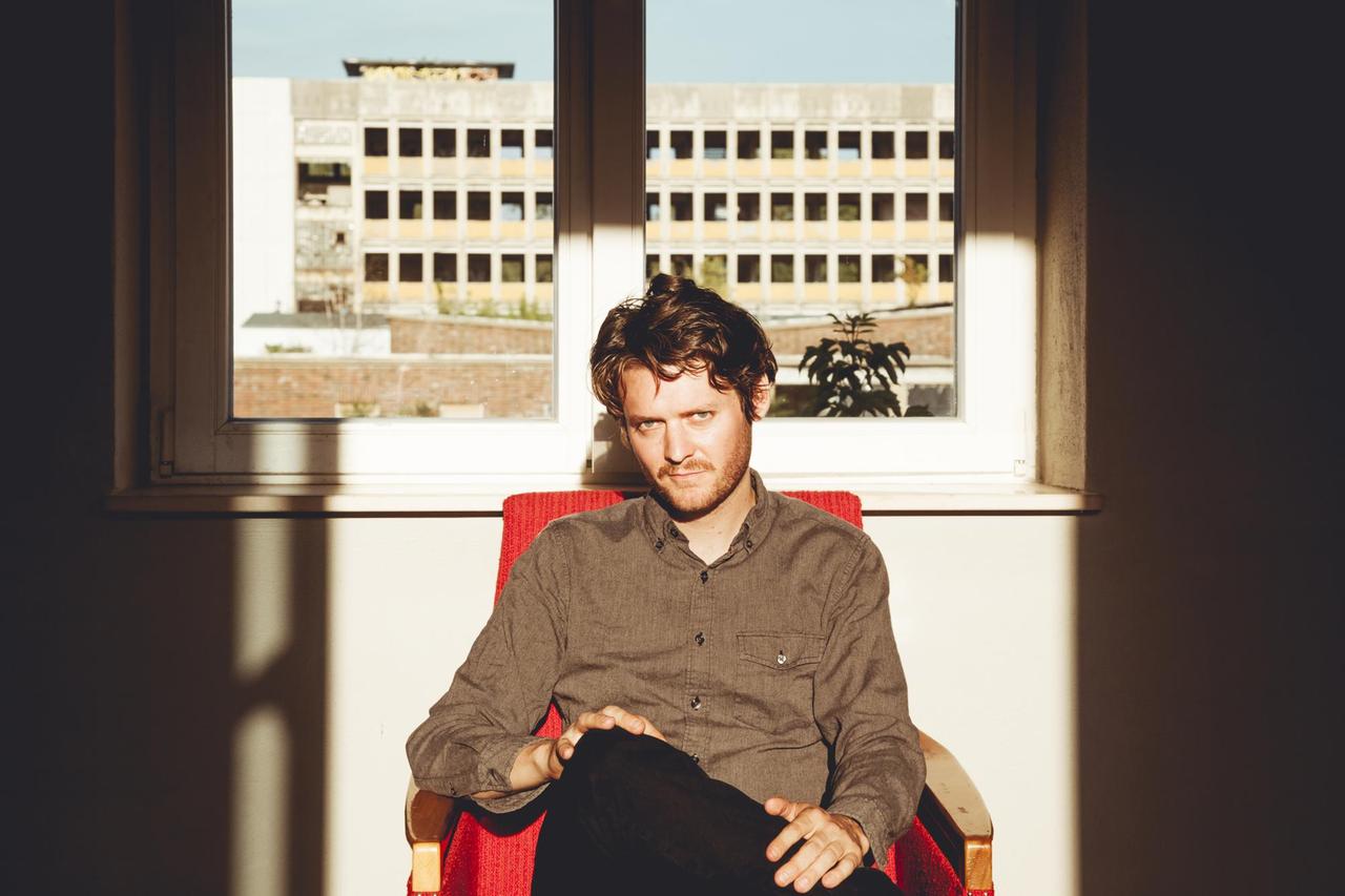 Zach Condon alias "Beirut" in Berlin 2018. Er sitzt auf einem rotem Sessel mit übergeschlagenen Beinen und schaut nachdenlick in die Kamera. Hinter ihm ein Fenster aus dem man auf ein leeres Gebäude schaut