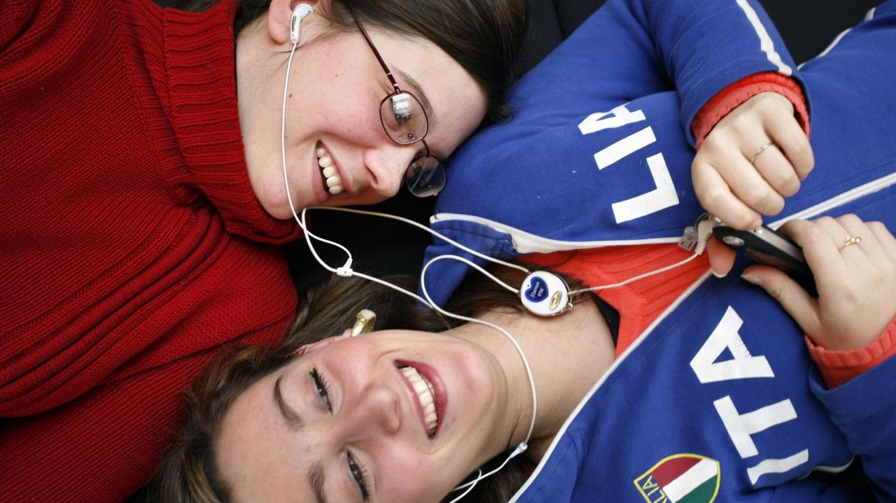 Zwei Frauen liegen Kopf an Kopf und teilen sich Ohrstöpsel. Eine trägt einen roten Pullover und Brille, die andere eine Trainingsjacke in blau mit der Aufschrift ITALIA.