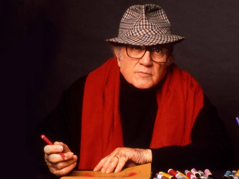 Der Regisseur Frederico Fellini mit Hut und rotem Schal skizziert etwas auf einem gelben mit Buntstiften.