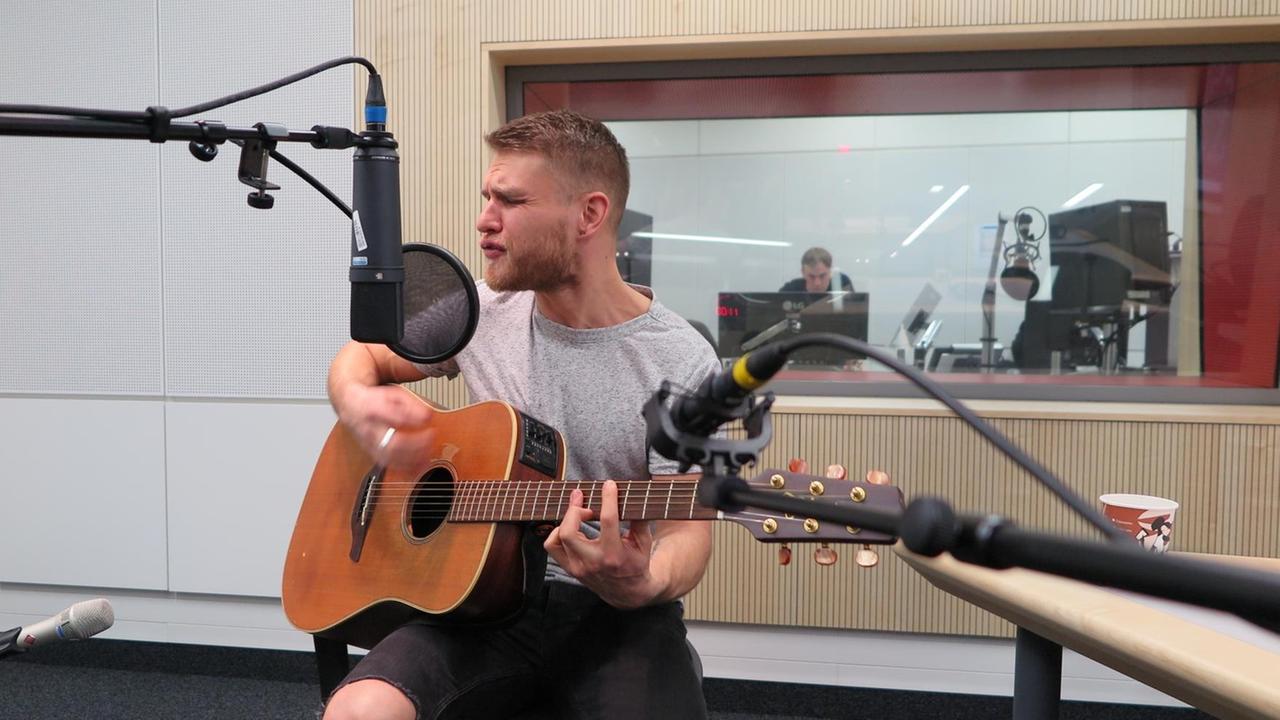 Ein Mann sitzt in einem Hörfunkstudio vor Mikrofonen, er spielt akustische Gitarre und singt mit geschlossenen Augen.