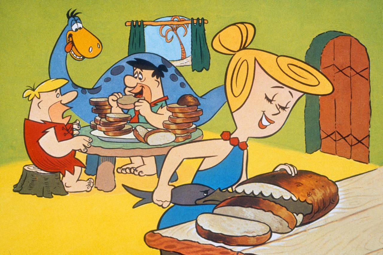 Szene aus der TV-Zeichentrickserie "Flintstones": Barney Rubble und Fred Flintstone unterhalten sich, Wilma Flintstone schneidet Brot mit einem Haifisch-Messer.