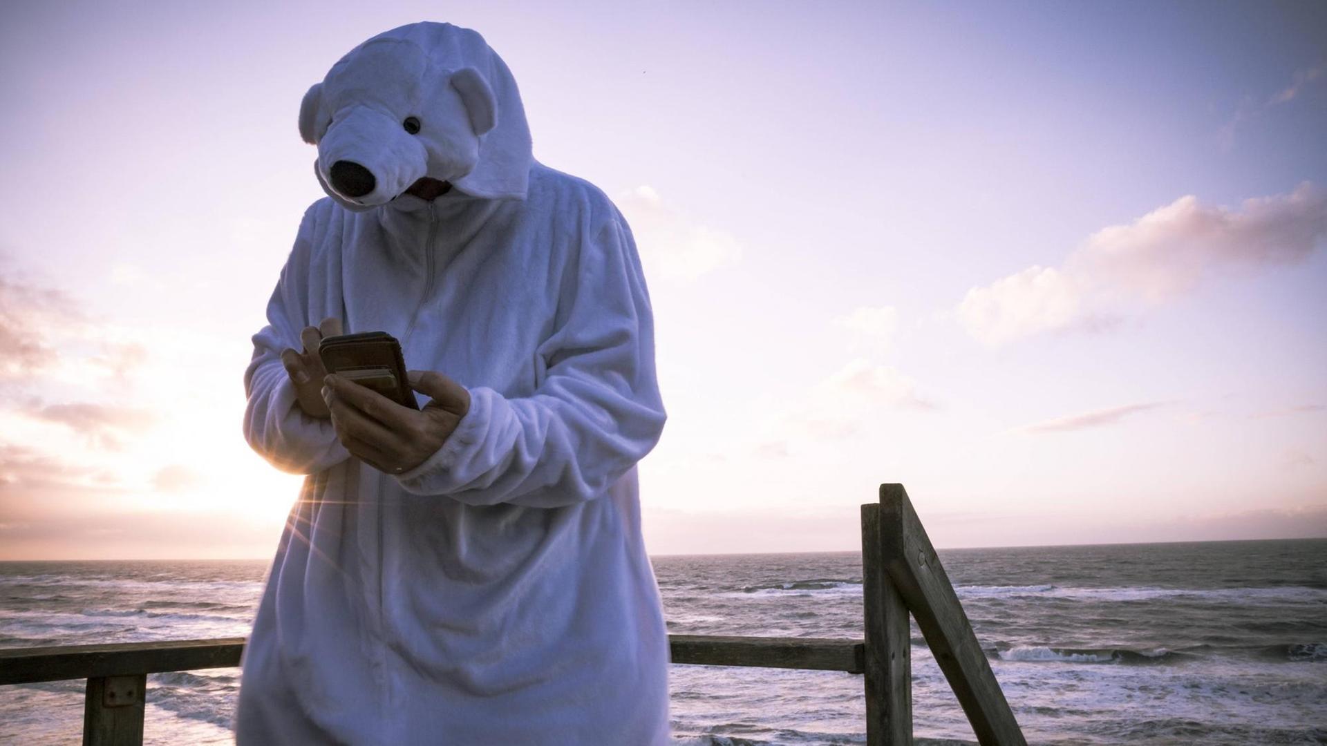 Ein Mann steht am Strand, trägt ein Eisbärkostüm und hält Smartphone in der Hand.