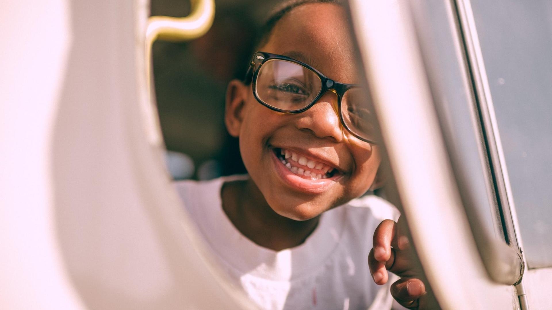 Ein grinsender Junge mit Brille schaut durch eine Art Luke oder Fenster