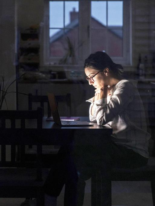 Eine Frau sitzt im dunkeln Raum an einem Tisch und arbeitet.