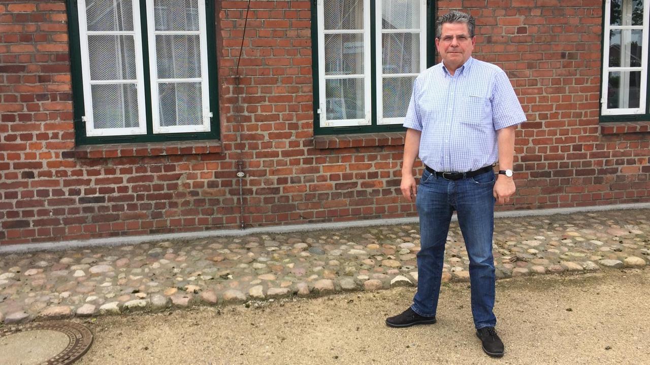 Ein Mann in einem hellen Hemd, dunklen Jeans und Brille steht vor einem Backsteinhaus. Der Mann heißt Walter Lorenzen, er ist stellvertretender Bürgermeister des Ortes Appen bei Hamburg.