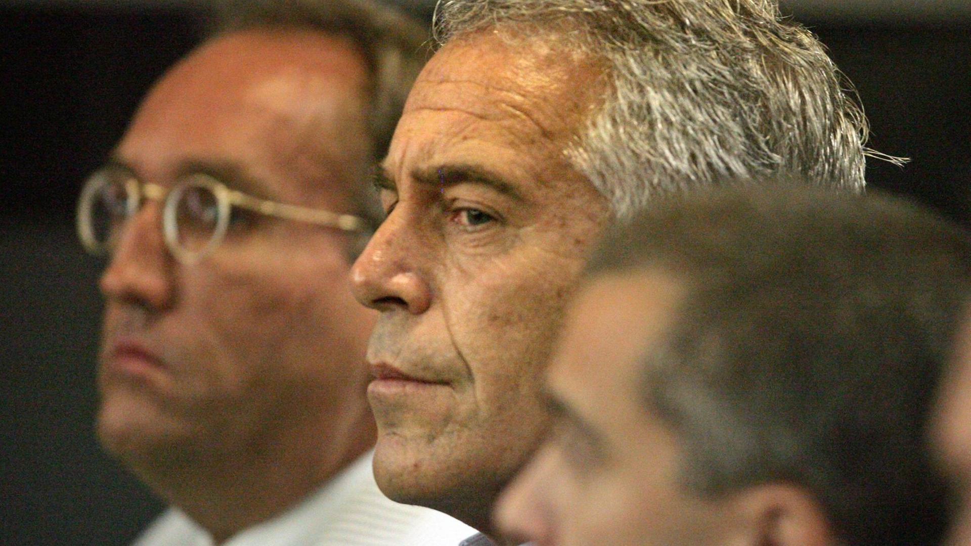 Jeffrey Epstein steht von der Seite gesehen zwischen zwei Männern.