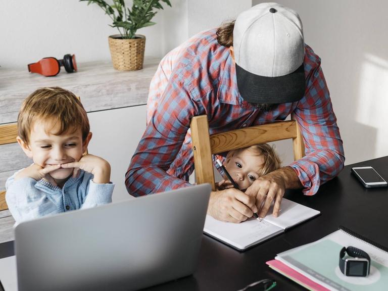Ein Vater sitzt mit seinen Kindern am Küchentisch über dem Laptop und den Lernaufgaben. (Symbolfoto)