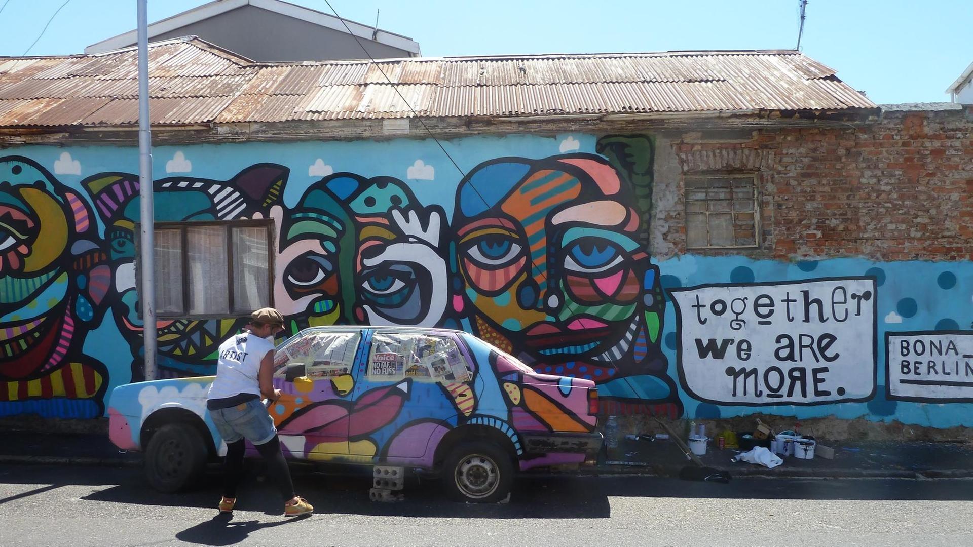 Ein buntes Graffiti aus Gesichtern an einer Häuserwand und über einem geparkten Auto. Rechts ebenfalls aufgesprayt der Spruch "Together We Are More".