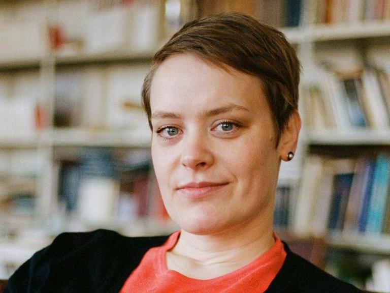 Hanna Engelmeier sitzt vor einer Bücherwand. Sie trägt ein hellrotes Oberteil und eine schwarze Jacke und schaut in die Kamera. Das Haar trägt sie kurz mit Seitenscheitel.