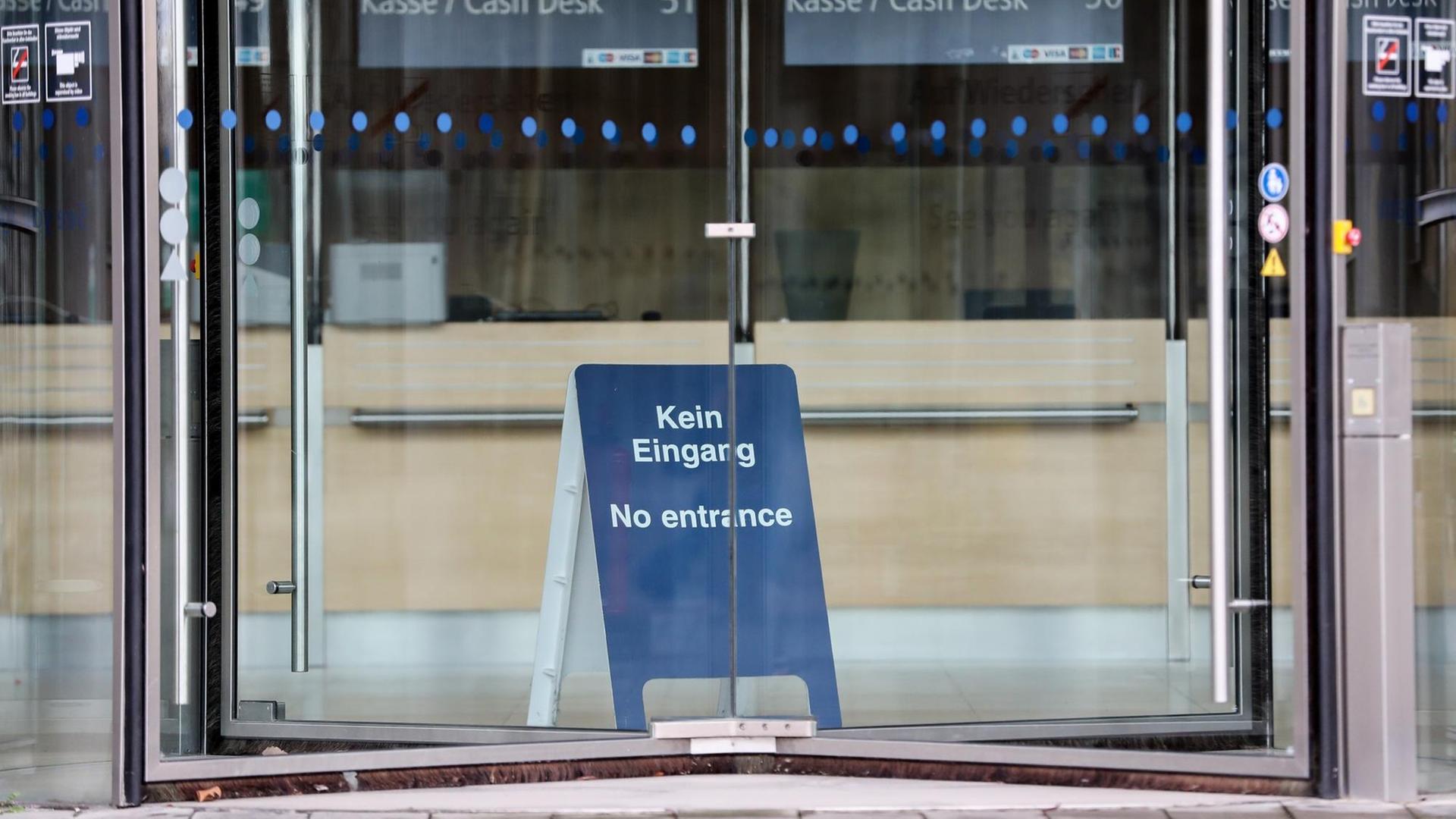 Durch eine Drehtür ist ein blaues Aufstellschild mit der Aufschrift 'Kein Eingang, No entrance' zu sehen. Im Hintergrund sind die leeren Kassenplätze.