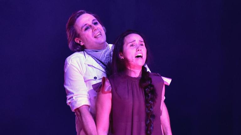 Zwei Frauen stehen direkt hintereinander mit geöffneten Mündern zum Singen. Sie werden rötlich-pink angeleuchtet und stehen vor einem dunkelblauen Hintergrund.