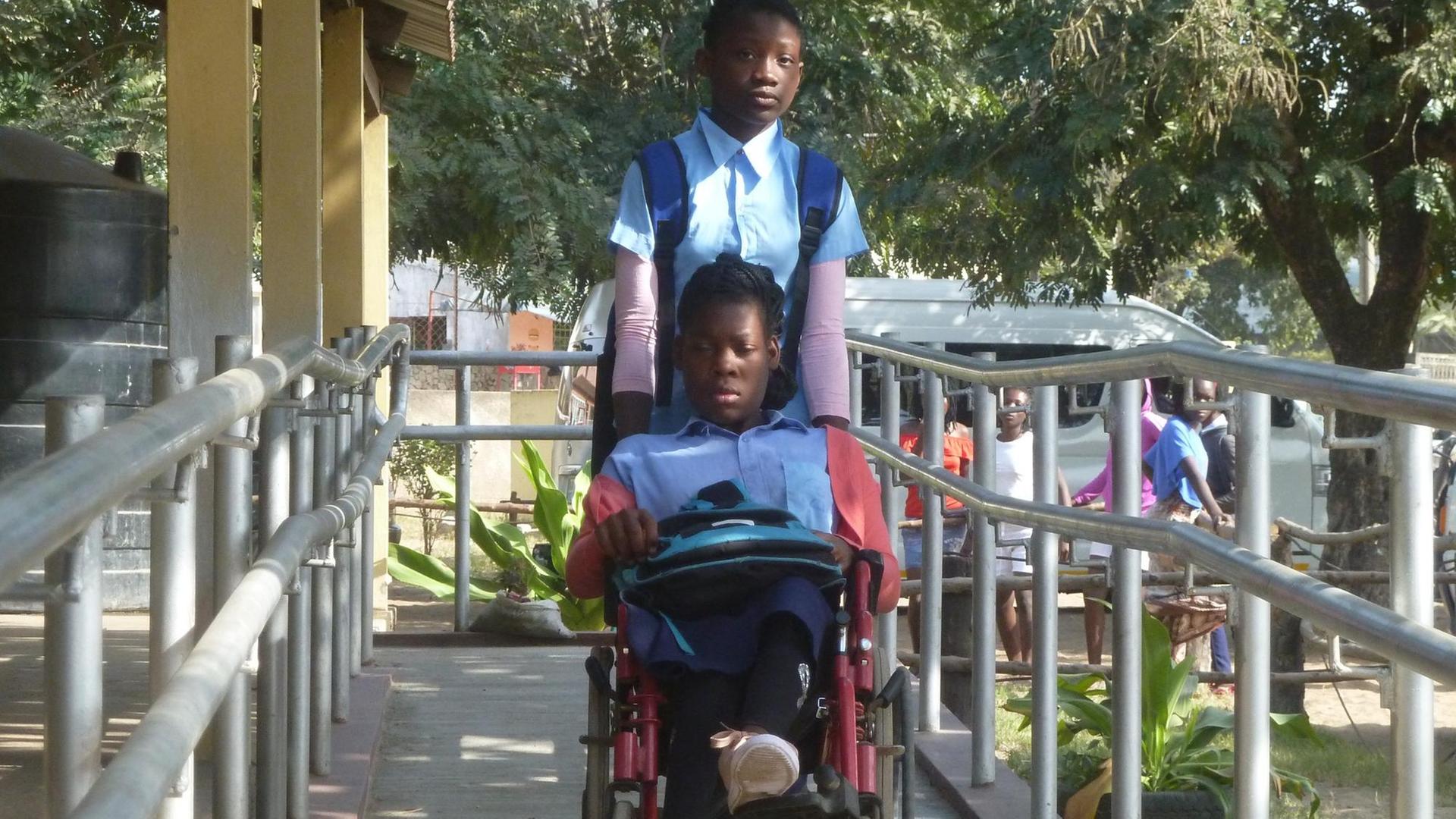 Das Bild zeigt ein Mädchen, das ein anderes Mädchen im Rollstuhl in die Schule bringt.
