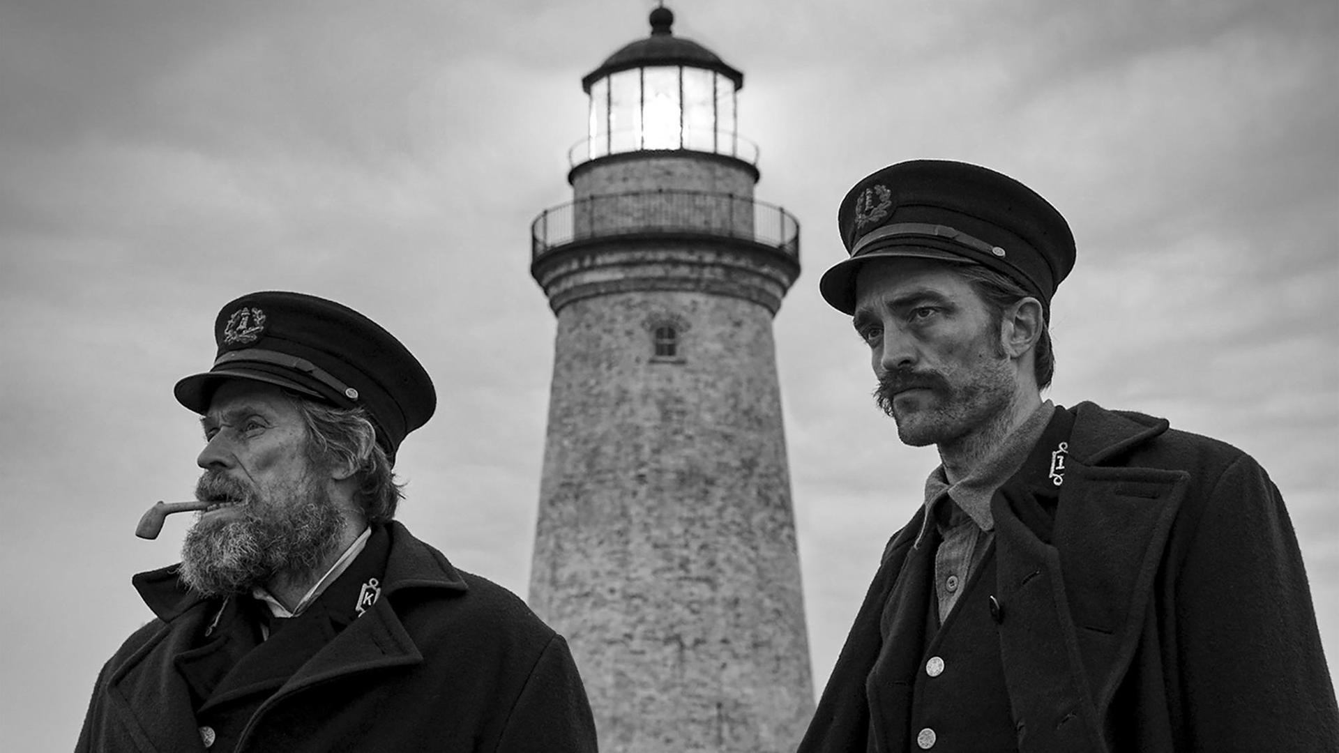 Die Schauspieler Willem Dafoe und Robert Pattinson spielen in "Der Leuchtturm" zwei Leuchtturmwärter am Ende des 19. Jahrhunderts. Die zwei Männer stehen in schwarz-weißer Kulisse vor einem Leuchtturm.