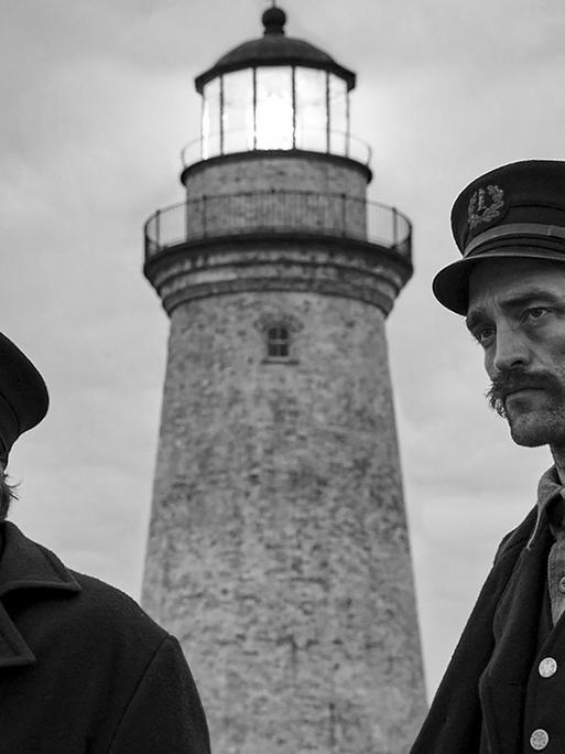 Die Schauspieler Willem Dafoe und Robert Pattinson spielen in "Der Leuchtturm" zwei Leuchtturmwärter am Ende des 19. Jahrhunderts. Die zwei Männer stehen in schwarz-weißer Kulisse vor einem Leuchtturm.