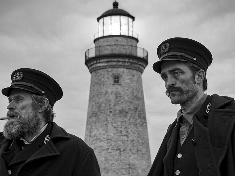 Die Schauspieler Willem Dafoe und Robert Pattinson spielen in "Der Leuchtturm" zwei Leuchtturmwärter am Ende des 19. Jahrhunderts. Die zwei Männer stehen in schwarz-weißer Kulisse vor einem Leuchtturm.