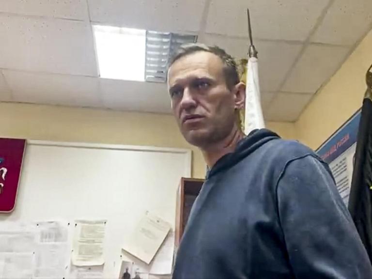 Nawalny im dunklblauen Pulli ist von unten fotografiert. Er steht in einem Dienstzimmer mit Fahnen und Wappen an der Wand und blickt ernst.