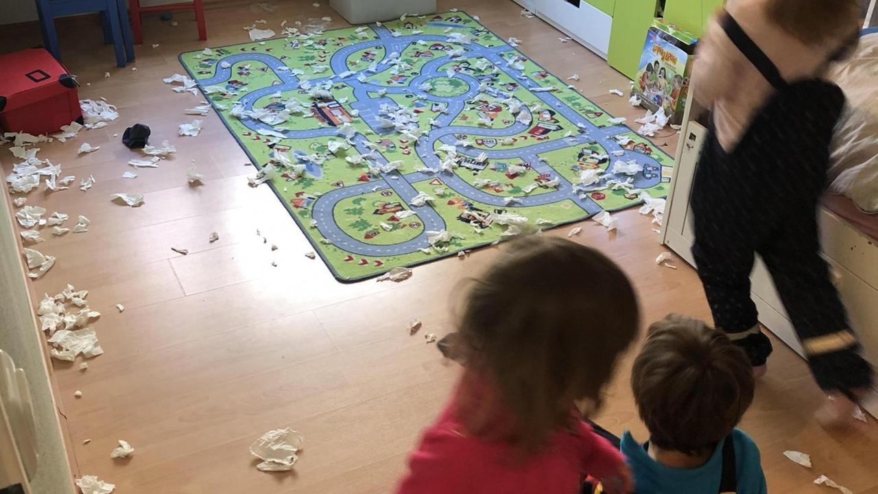 Der Fußboden des Kinderzimmers ist mit einem Spielteppich und vielen Papierschnipseln bedeckt. Zu sehen sind unscharf drei kleine Kinder.