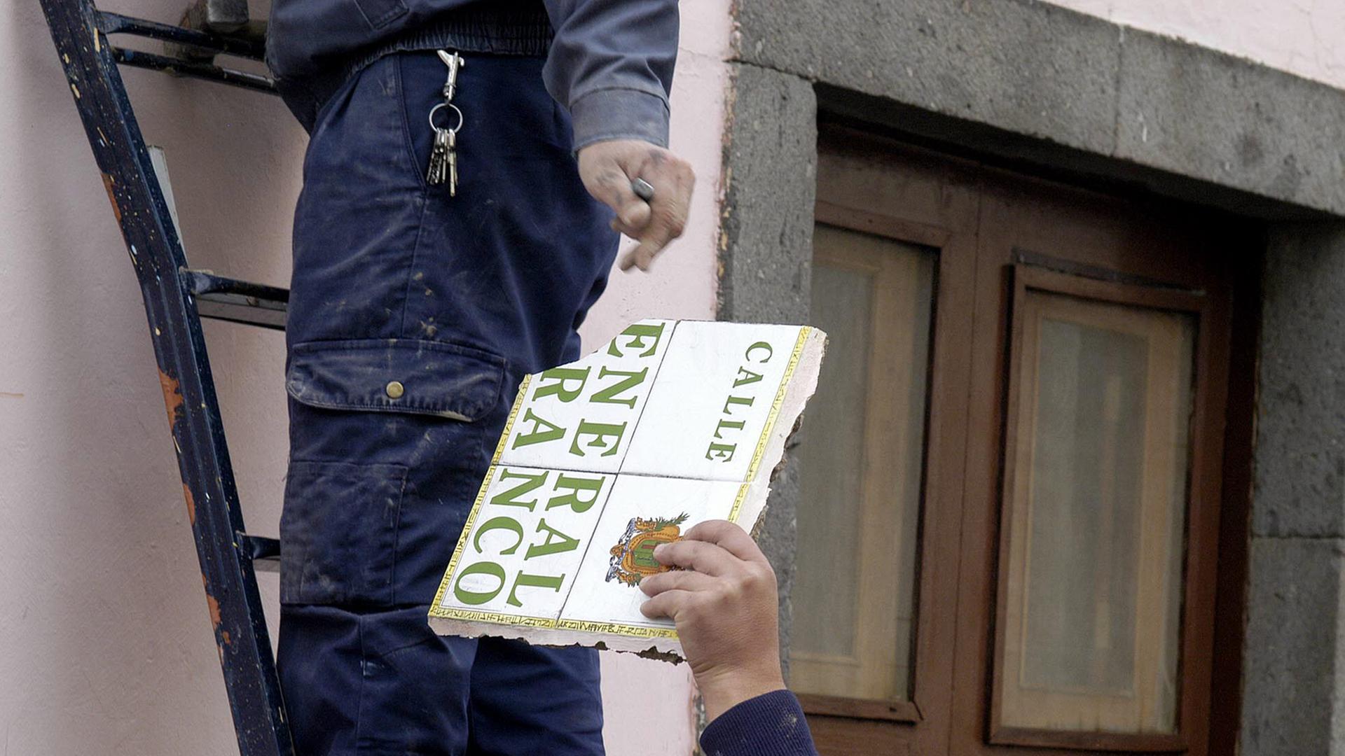 Arbeiter wechseln ein Straßenschild aus, auf dem "Calle General Franco" steht, ein Arbeiter steht auf einer Leiter, nur seine Beine sind sichtbar, ein anderer nimmt das Schild in die Hand