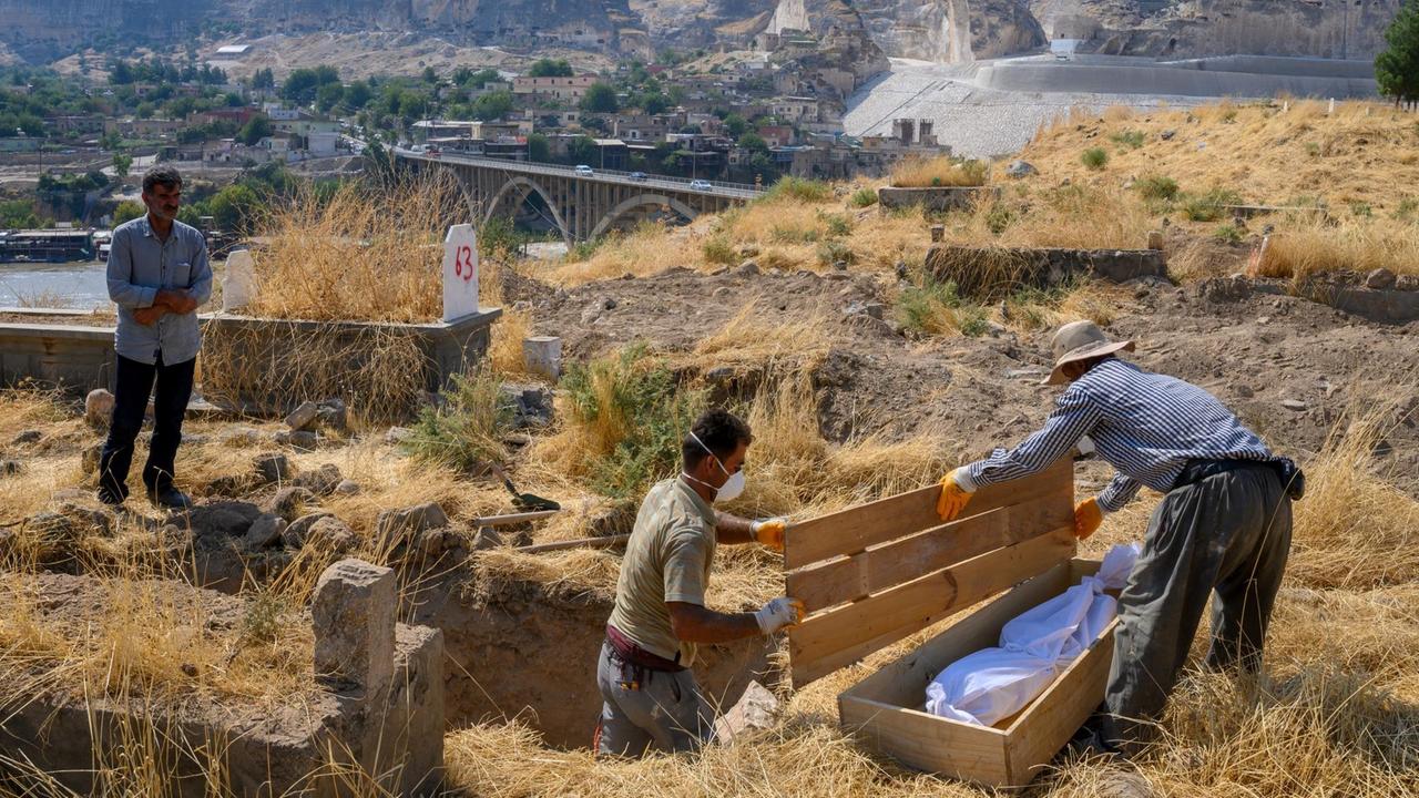 Zwei Männer holen einen in weißen Tüchern eingewickelten Leichnahm aus einem Grab und legen ihn in einen Holzsarg. Ein weiterer Mann sieht zu. Im Hintergrund ist die Stadt Hasankeyf zu erkennen.