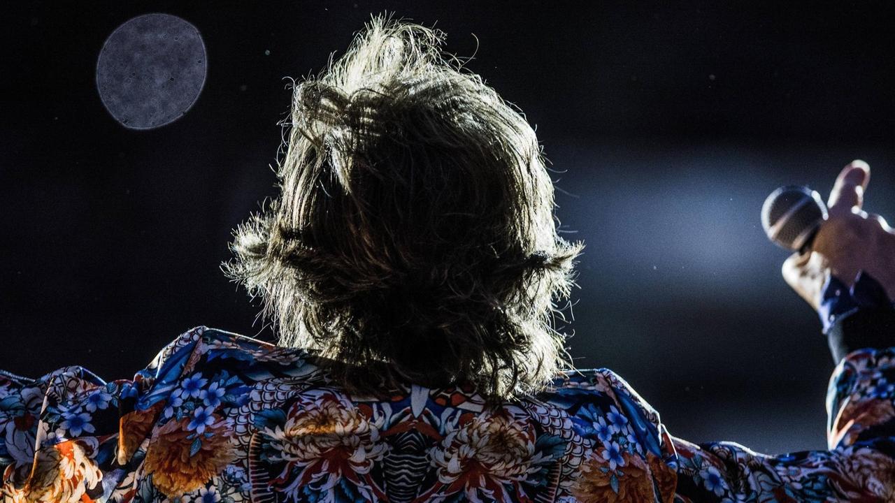 Mick Jagger steht mit dem Rücken zur Kamera. Das Licht hebt seine Haare hervor. Sein Hemd ist bunt. Er macht eine typische Rock-Pose.